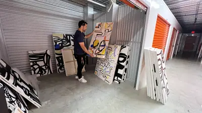 Inside the storage unit: canvases stacked and leaning along a corridor of orange roll-doors, one being lifted clear.