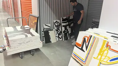 A trolley stacked with canvases in the storage corridor, more propped against the wall behind, one held up to the light.